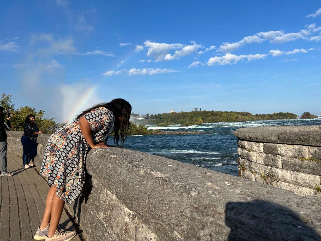 A person leans over a stone railing beside a river, with a visible rainbow in the mist. Another person stands nearby, in a sunny scene with blue skies.