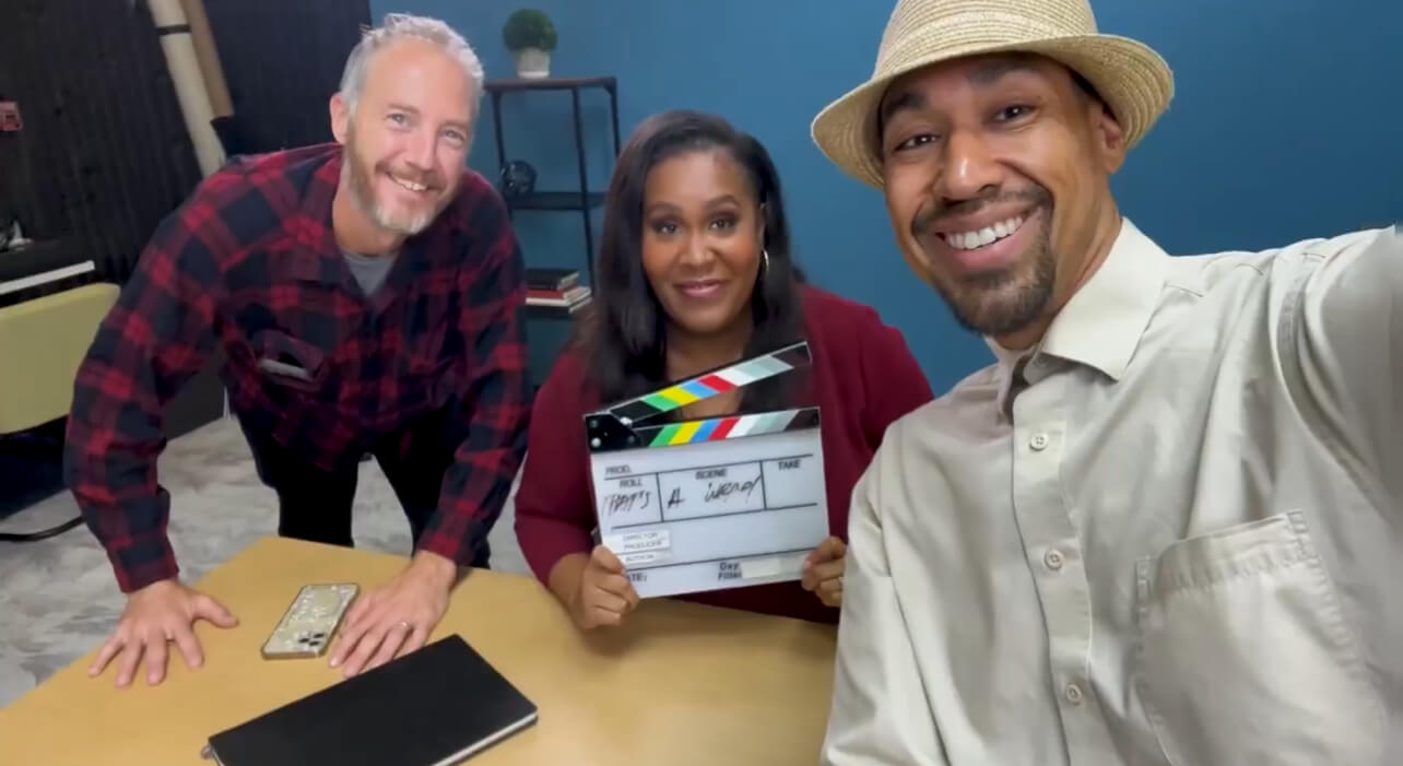 Three people are smiling at the camera, one holding a film slate. They're in a room with a blue wall and modern furniture.